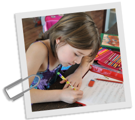 Auf dem Bild sieht man eine Grundschlerin ber ein Blatt Papier gelehnt. Sie schreibt mit Bleistift darauf. Daneben liegen Bundstifte in einer Federmappe.